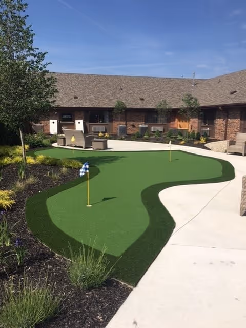 Outdoor courtyard area at Homeview Health and Rehabilitation Center featuring a small artificial putting green with two golf holes, surrounded by landscaped plants and trees, with a brick building in the background under a clear blue sky.