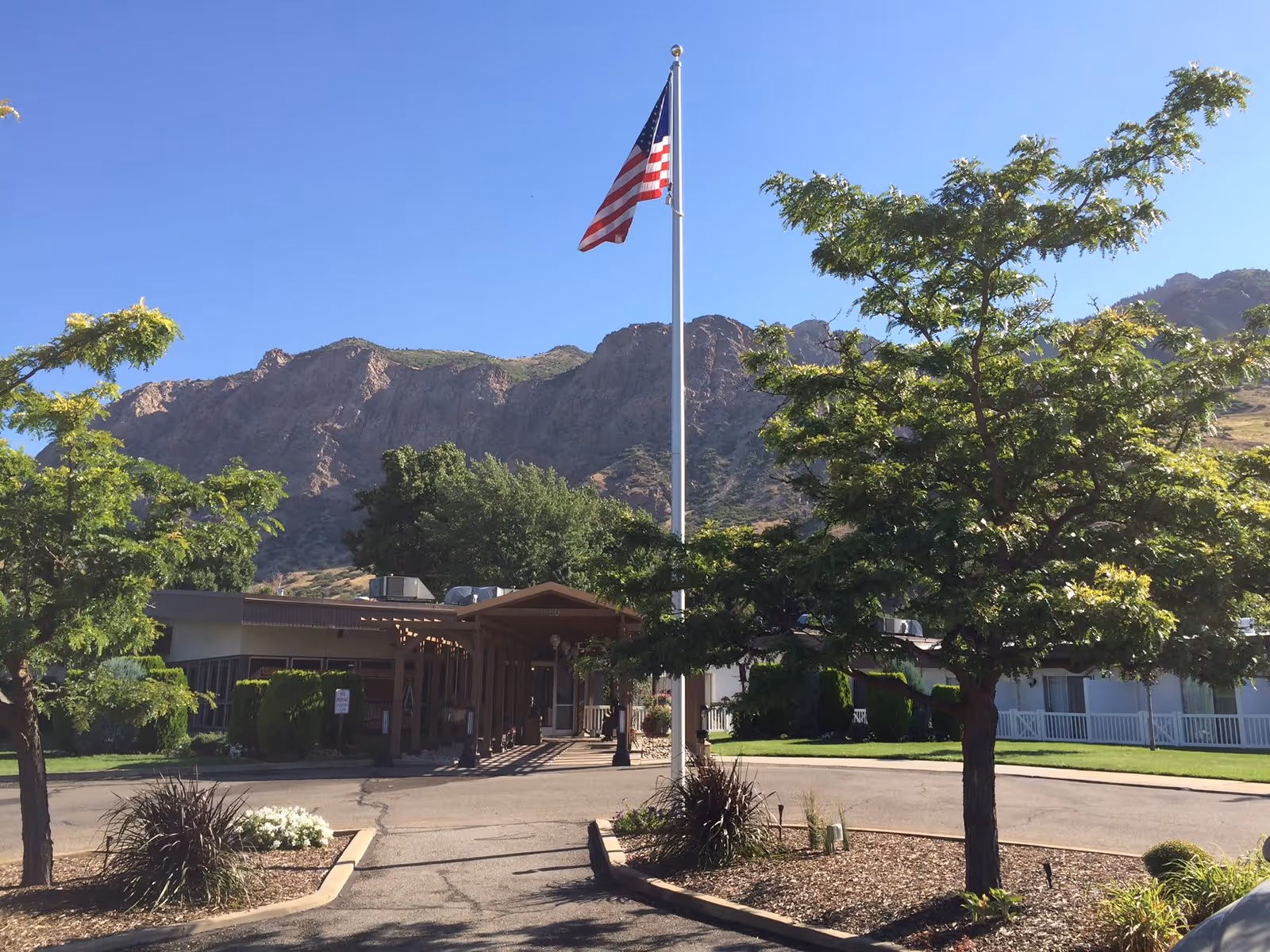 Entrance of Gardens Assisted Living facility with a flagpole displaying the American flag in the center. The building is surrounded by trees and landscaping, with mountains visible in the background under a clear blue sky.