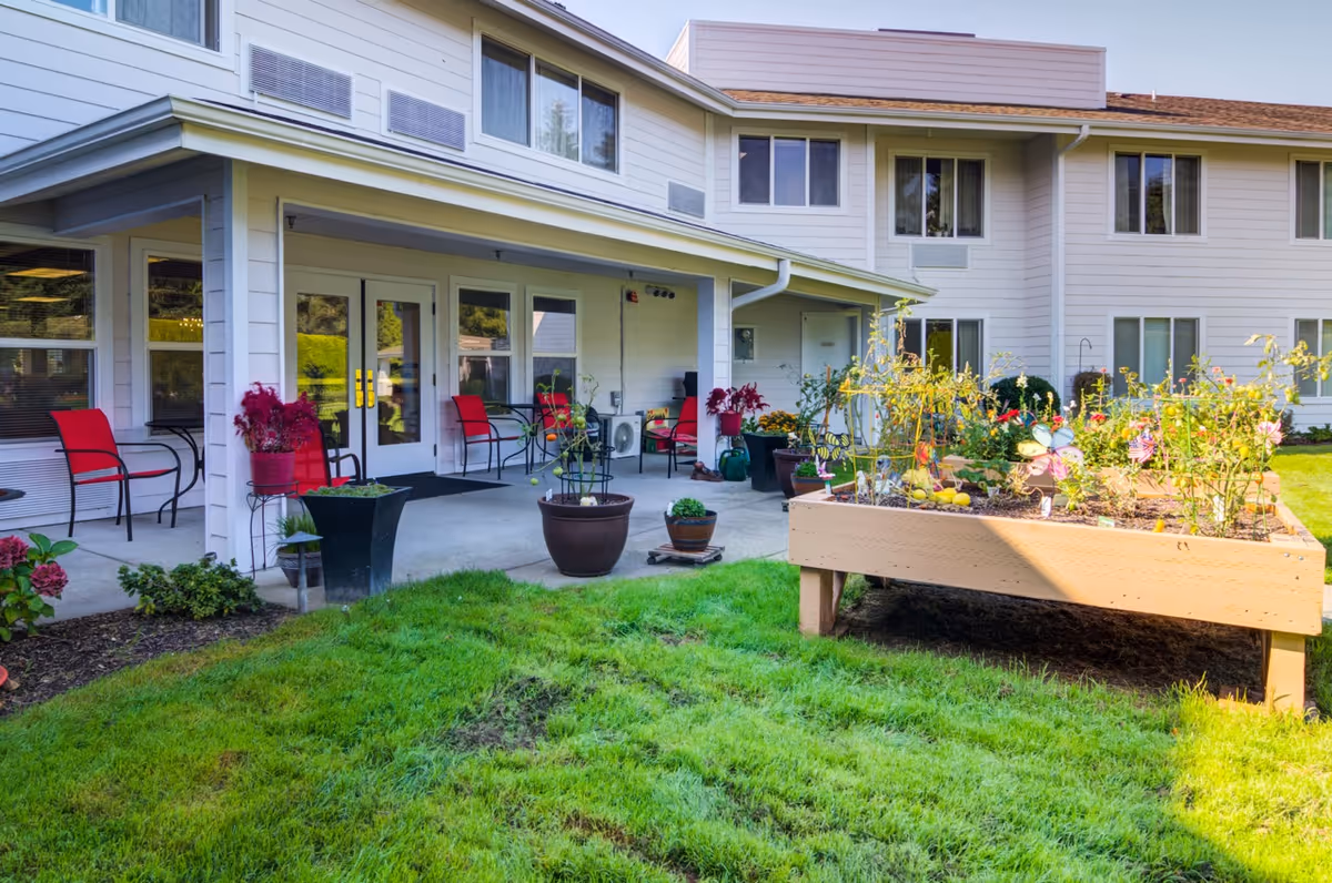 Outdoor patio area of a senior living facility with red chairs, potted plants, and a raised garden bed with various plants and flowers. The building has white siding and multiple windows.