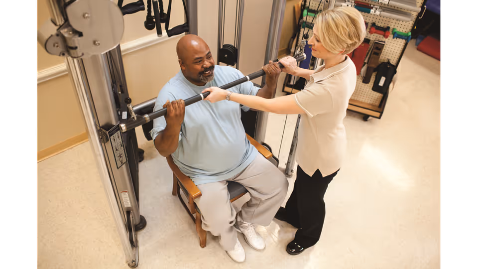 A man seated on a chair using a weight machine for exercise while a woman, possibly a physical therapist or trainer, assists and supports him by holding the exercise bar.