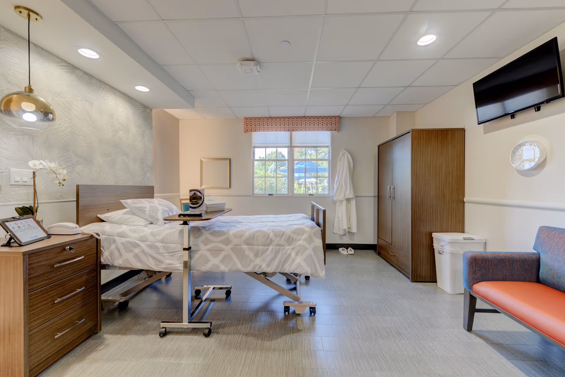 Well-lit senior care patient room with an adjustable bed, bedside table, wardrobe, wall-mounted TV and seating.