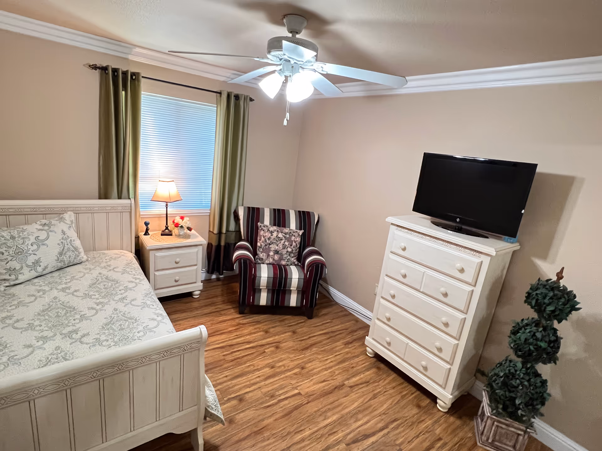 Bright bedroom with bed, nightstand, striped chair, dresser, TV, and ceiling fan.