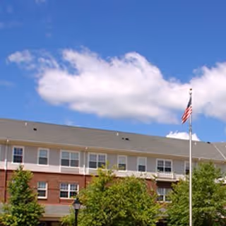 Two-story brick senior living building with a flagpole and trees against a blue sky.