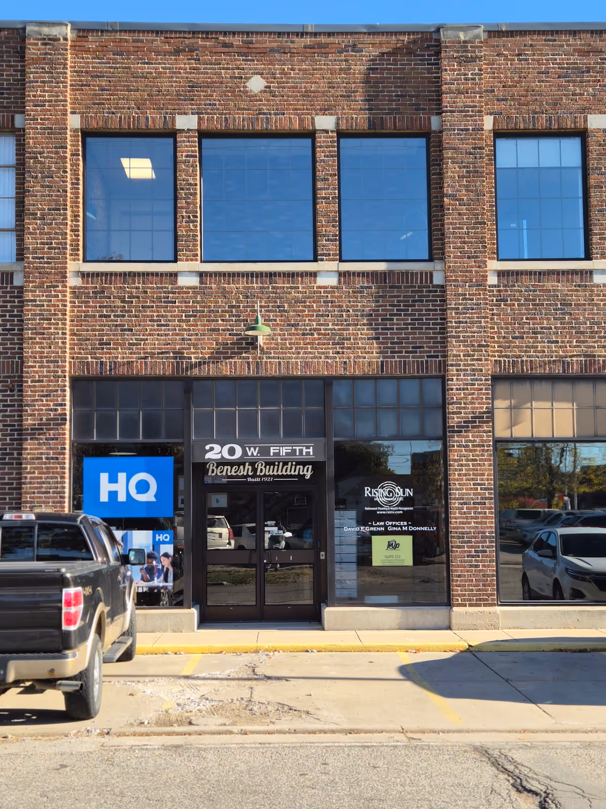 Brick storefront building with large windows, glass doors, signage, and parked vehicles in front.