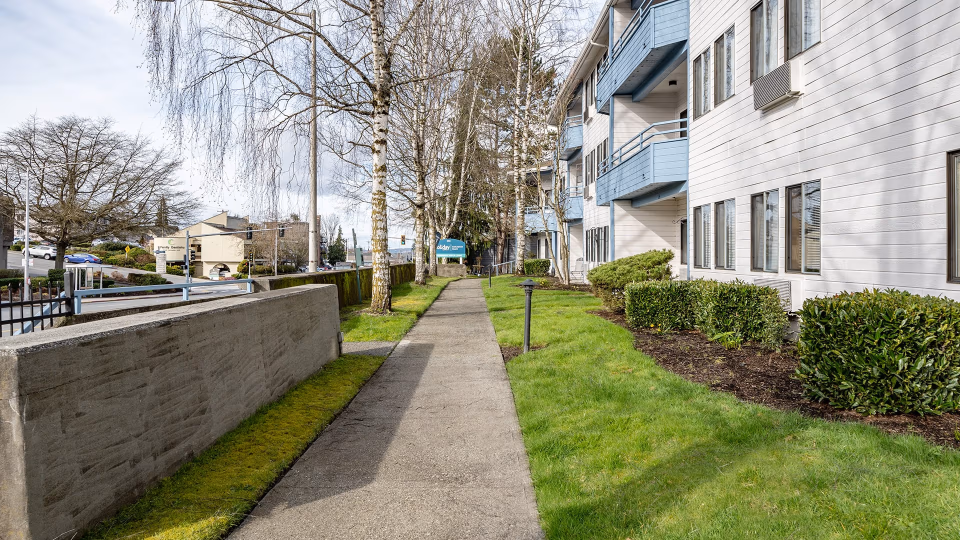 Sidewalk running alongside a multi-story residential building with blue balconies and white siding. The walkway is bordered by a low concrete wall on one side and a grassy area with shrubs and trees on the other. The sky is partly cloudy.