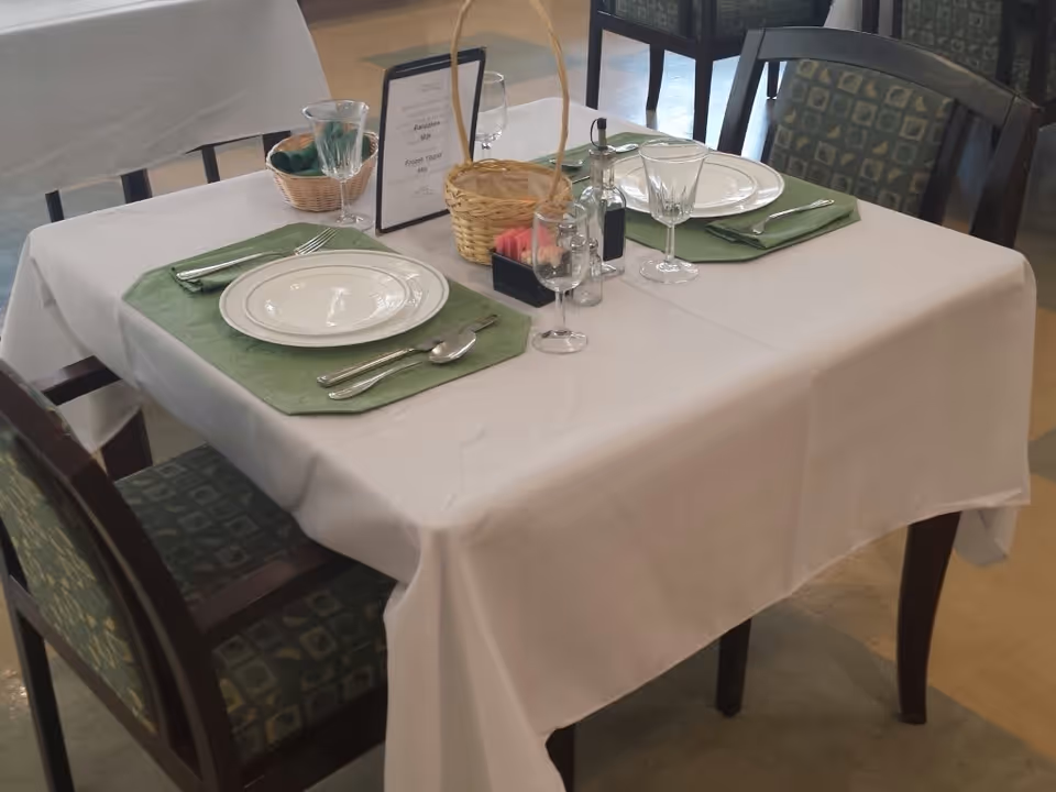 A dining table set for two with plates, wine glasses, utensils, green placemats and a white tablecloth in a communal dining area.