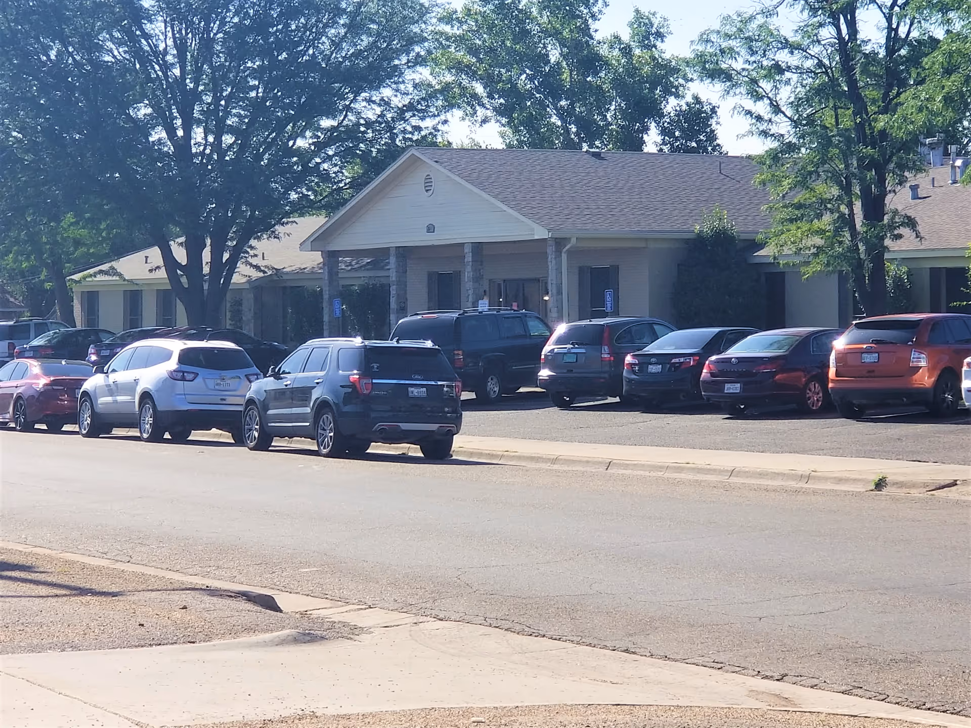 Exterior view of Georgia Manor Nursing Home building with a parking lot in front filled with several parked cars. The building has a covered entrance supported by stone columns and is surrounded by trees.