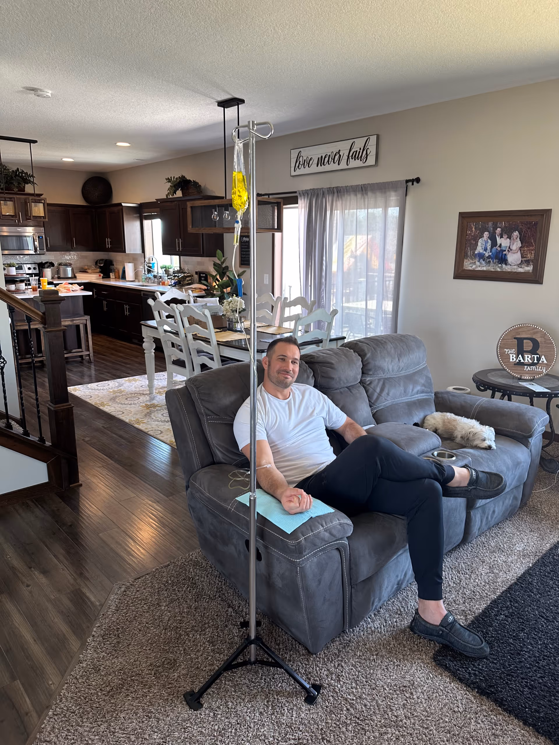 Man lounging on a gray sofa with IV pole in an open living area and kitchen.