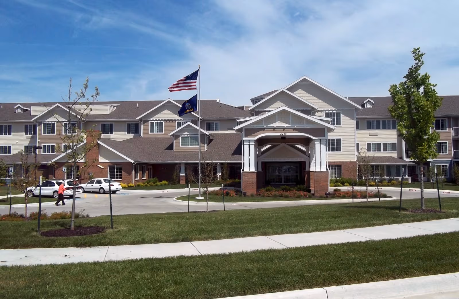 Front exterior of a multi-story senior living facility with a covered entrance, flagpoles, lawn, and parked cars.