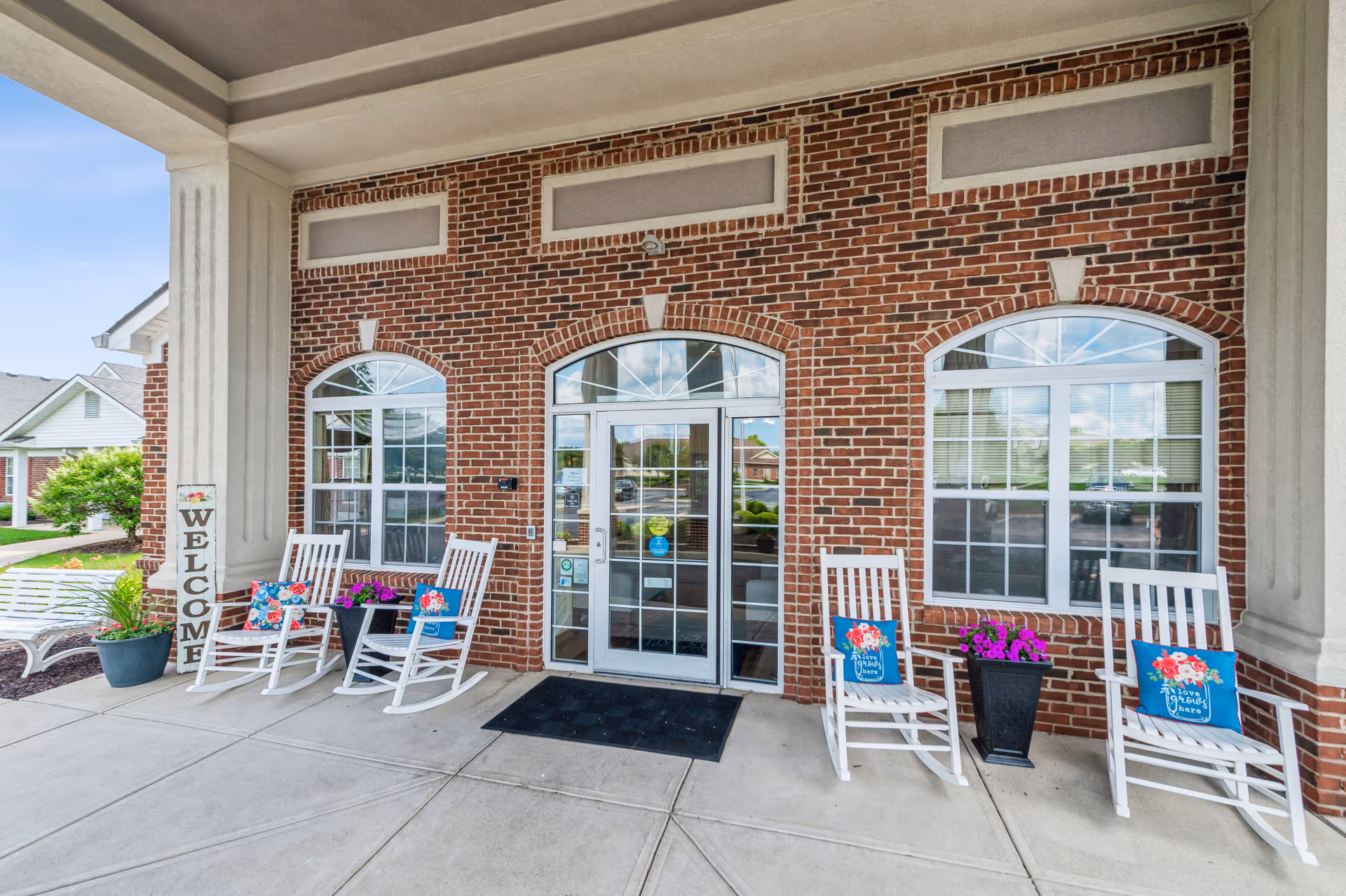 Entrance area of a brick building with a covered porch featuring four white rocking chairs with colorful floral cushions, two black planters with purple flowers, a vertical wooden welcome sign, and a glass door with windows on either side.