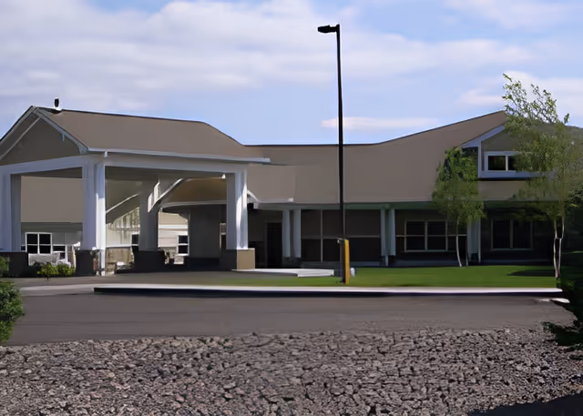 Exterior view of a single-story building with a covered entrance supported by white columns, a lamp post in front, and some trees and grass around the building under a partly cloudy sky.