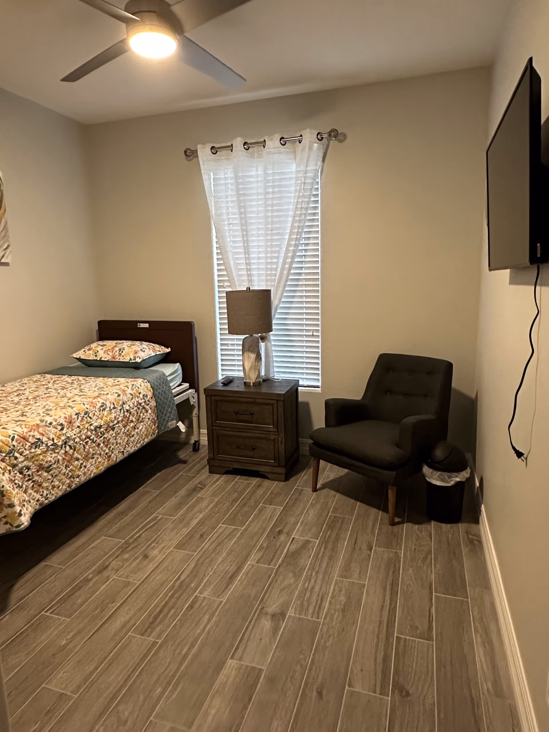 A small bedroom with a single bed, nightstand, chair, lamp, and wall-mounted TV.