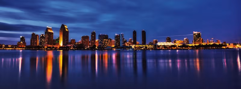 Panoramic nighttime city skyline with illuminated buildings reflecting across calm water.