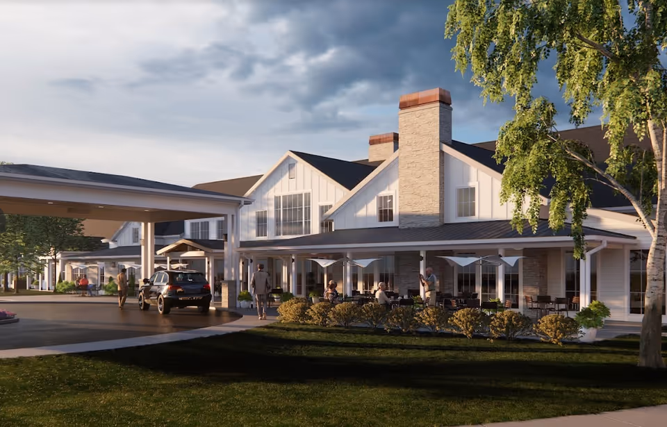 Exterior view of Cedarhurst Senior Living of Topeka showing a large building with white siding and stone chimneys. There is a covered driveway with a car and people walking nearby. Outdoor seating with umbrellas and people sitting is visible under a porch. Trees and bushes surround the area under a partly cloudy sky.