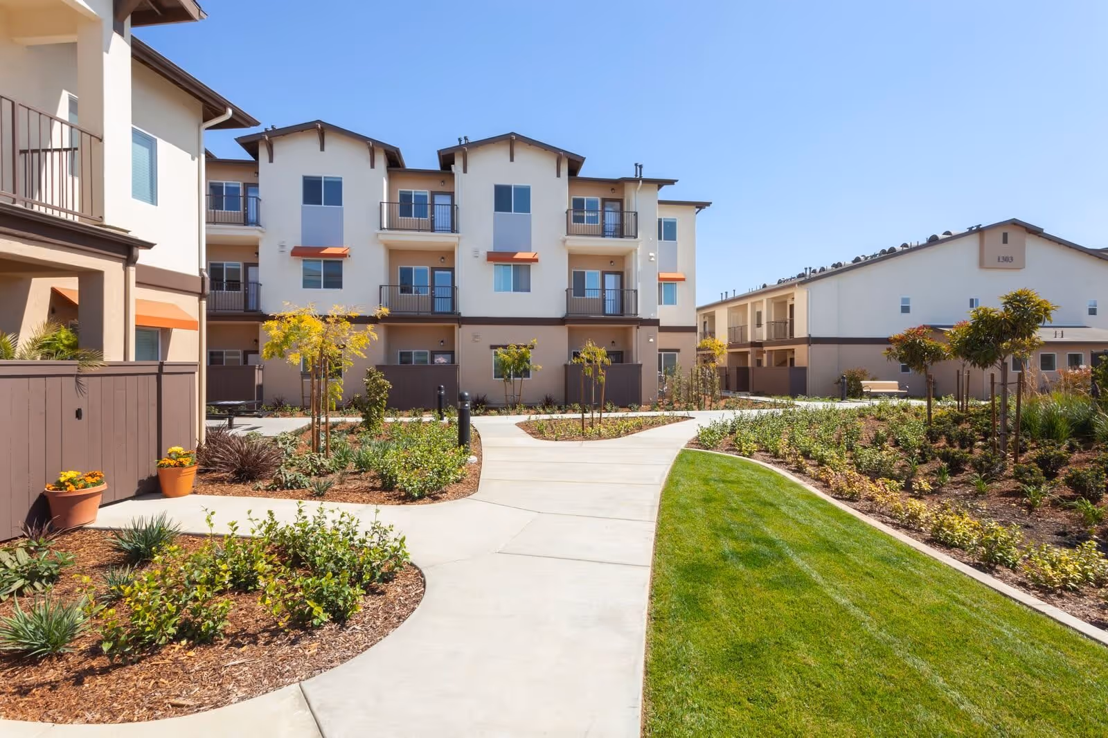 Courtyard with a paved walkway, manicured lawns and planted beds in front of multi-story apartment buildings under a clear blue sky.