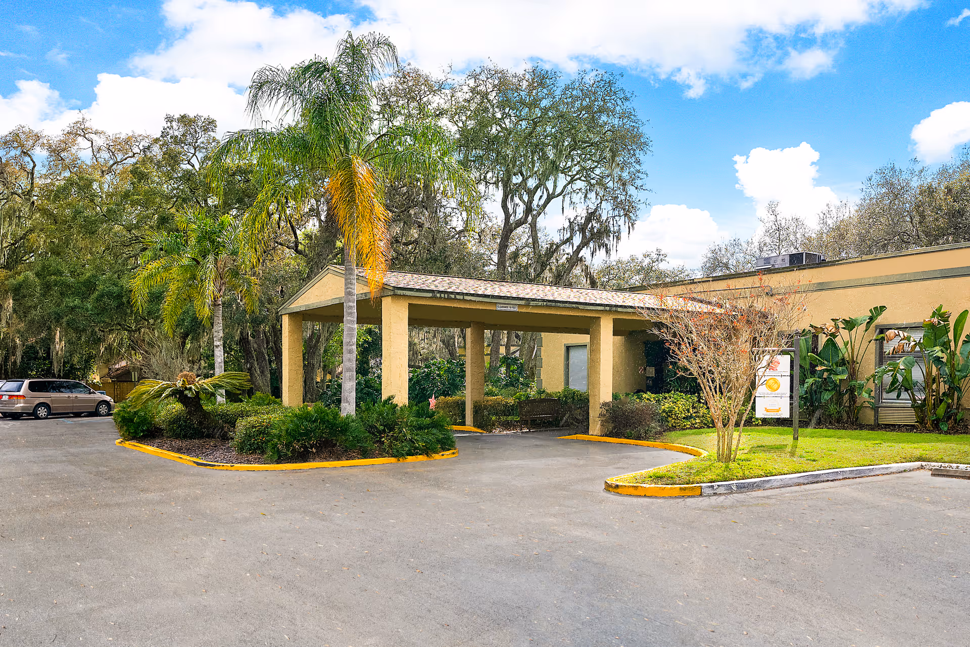 Entrance area of Fairway Oaks Center with a covered drop-off zone surrounded by palm trees and other greenery. The building is single-story with a beige exterior. There is a parking area with a vehicle visible on the left side and a partly cloudy blue sky above.