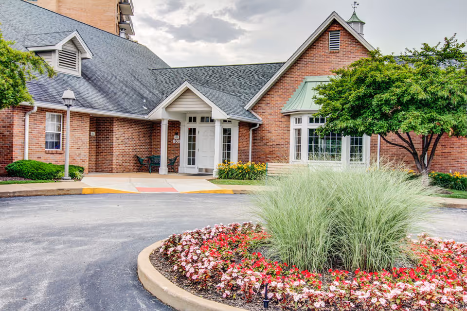 Front entrance of a brick senior living building with a covered doorway, circular flowerbed with ornamental grass, and a driveway.