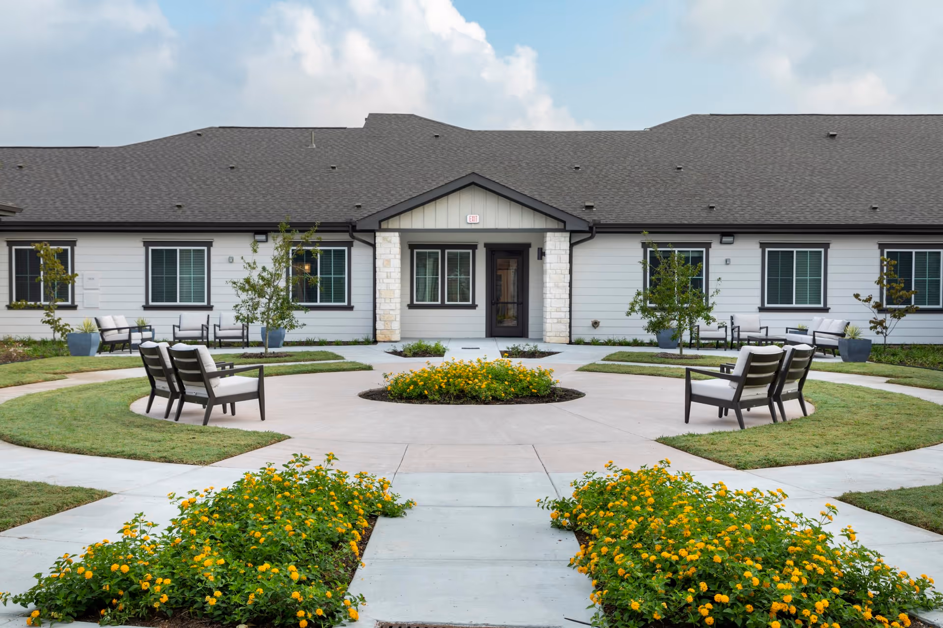 Front exterior of a single-story senior living building with a circular walkway, benches, and yellow flowerbeds.