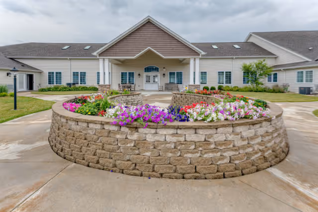 Front exterior view of a single-story senior living facility with beige siding and a gabled roof. In front of the building is a circular raised flower bed made of stone bricks, filled with colorful flowers including purple, white, red, and pink blooms. The entrance has double doors with windows on either side and a covered porch area.