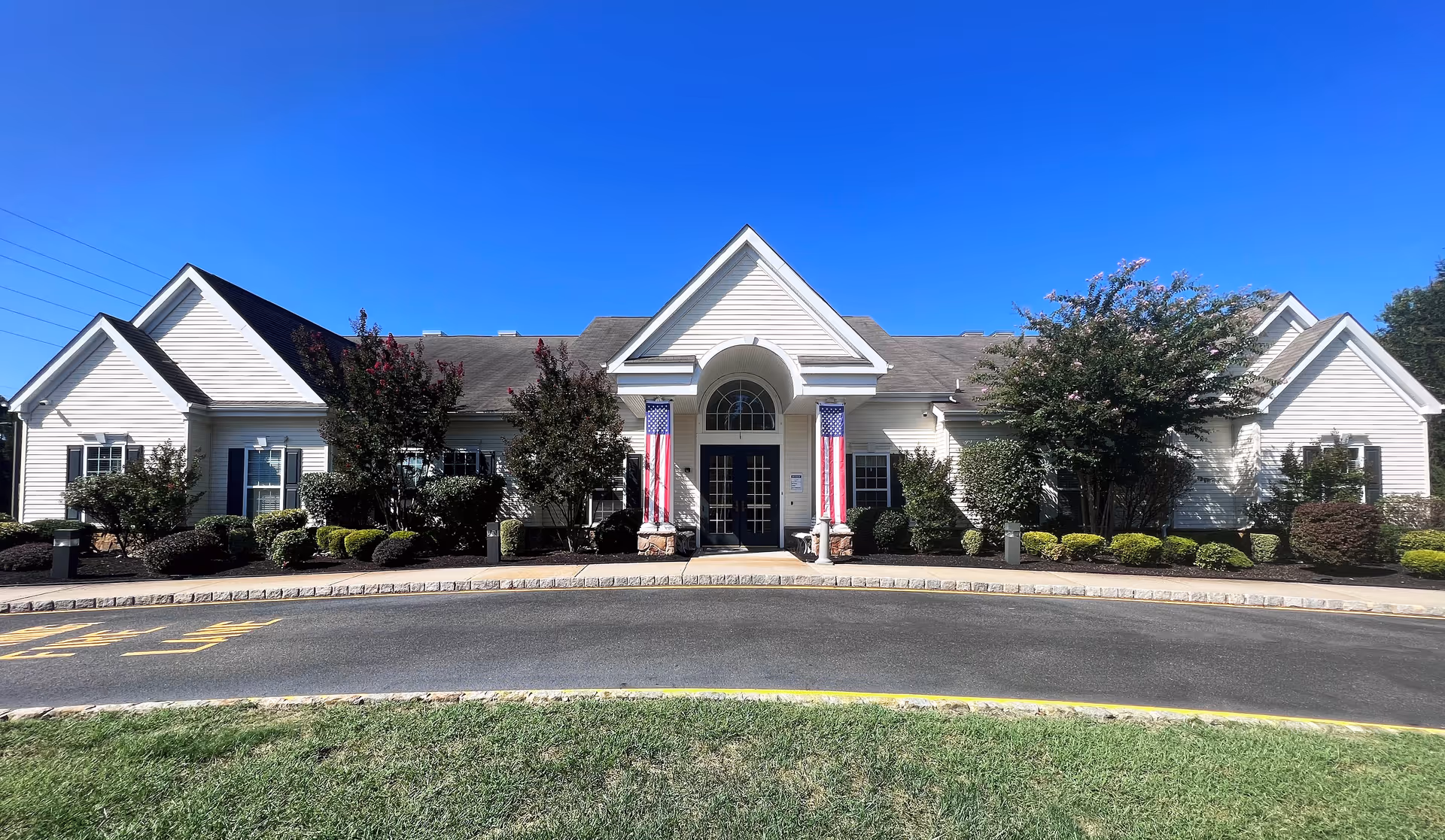 Single-story white building with a central entrance flanked by American flags, landscaped shrubs, and a curved driveway under a clear blue sky.