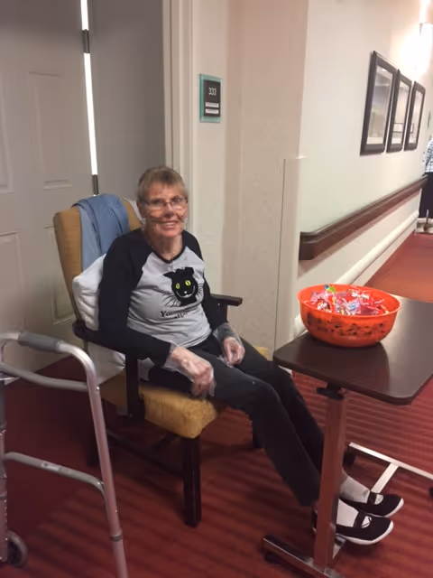 An elderly woman sitting in a chair in a hallway of a senior living facility. She is wearing a black and gray shirt with a cat graphic, black pants, and black shoes with white socks. She has gloves on her hands and is smiling. A walker is positioned next to her chair, and a table with a large orange bowl filled with candy is in front of her. The hallway has red carpet and framed pictures on the wall.