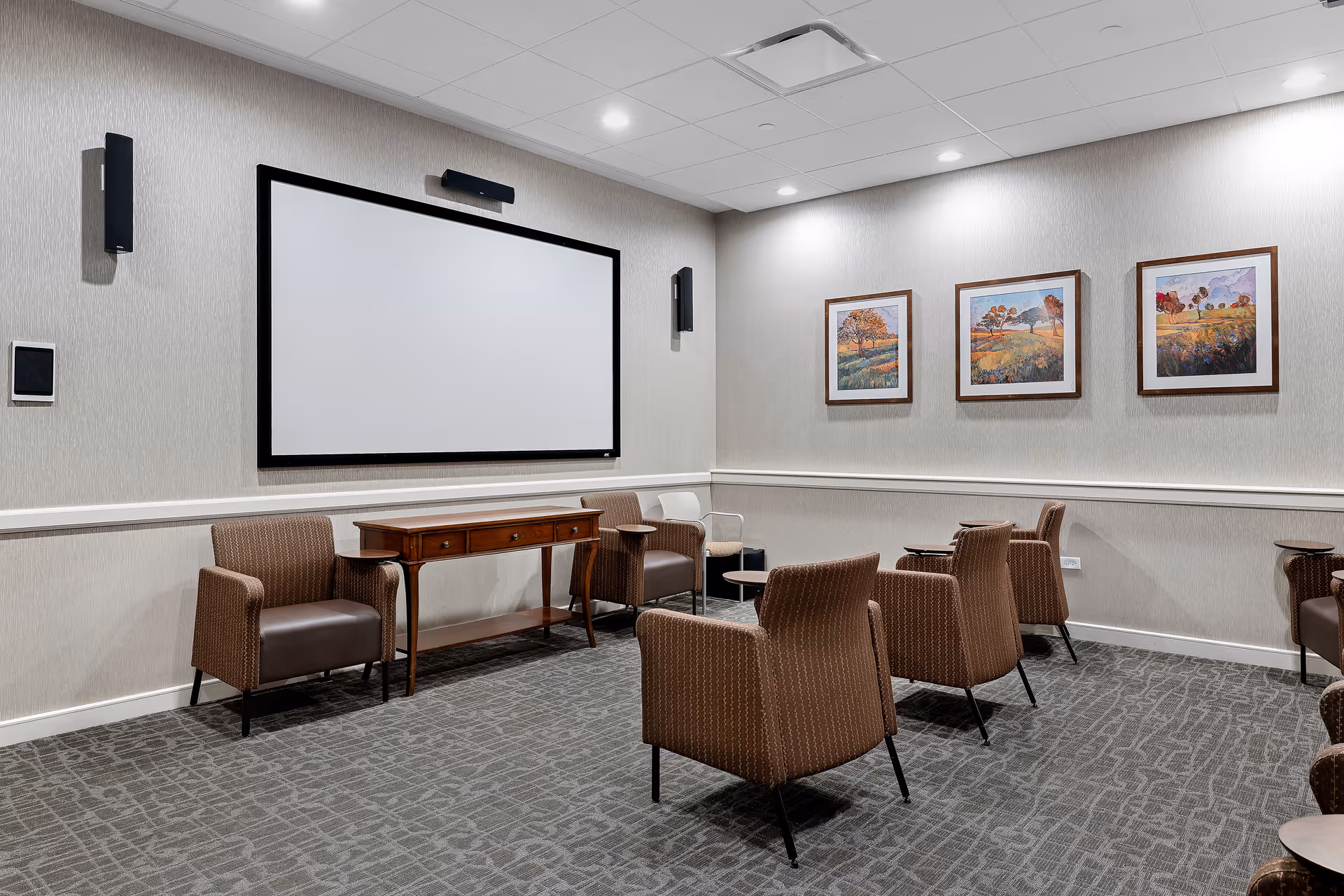 Small screening room with a large blank projection screen, several upholstered chairs, a wooden console table, and three framed landscape paintings on the wall.
