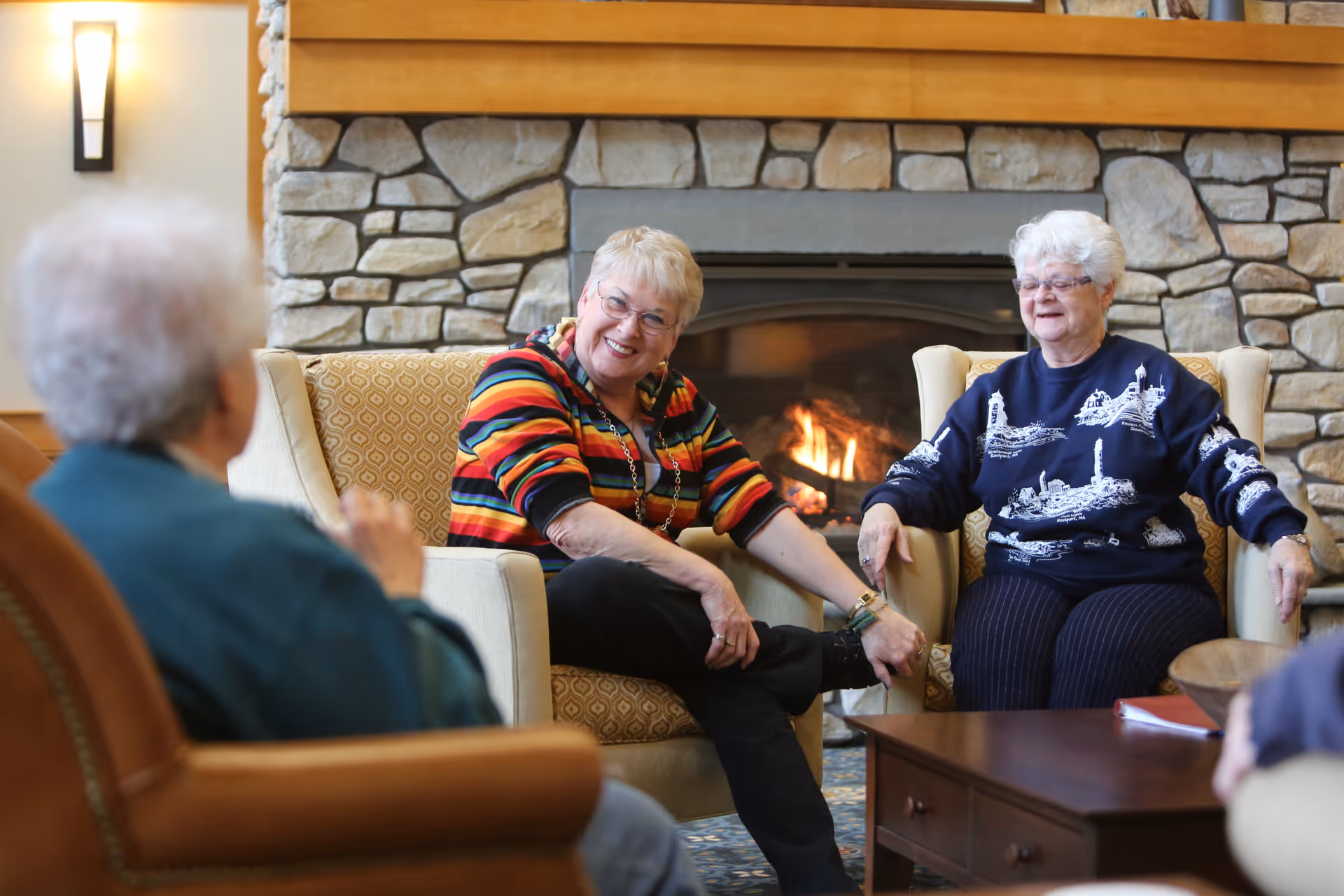 Three elderly women sitting and chatting in a cozy living room with a stone fireplace in the background. Two women are seated in armchairs facing the camera, smiling and engaging in conversation, while the third woman is partially visible from behind. The room has warm lighting and a wooden coffee table in front of them.