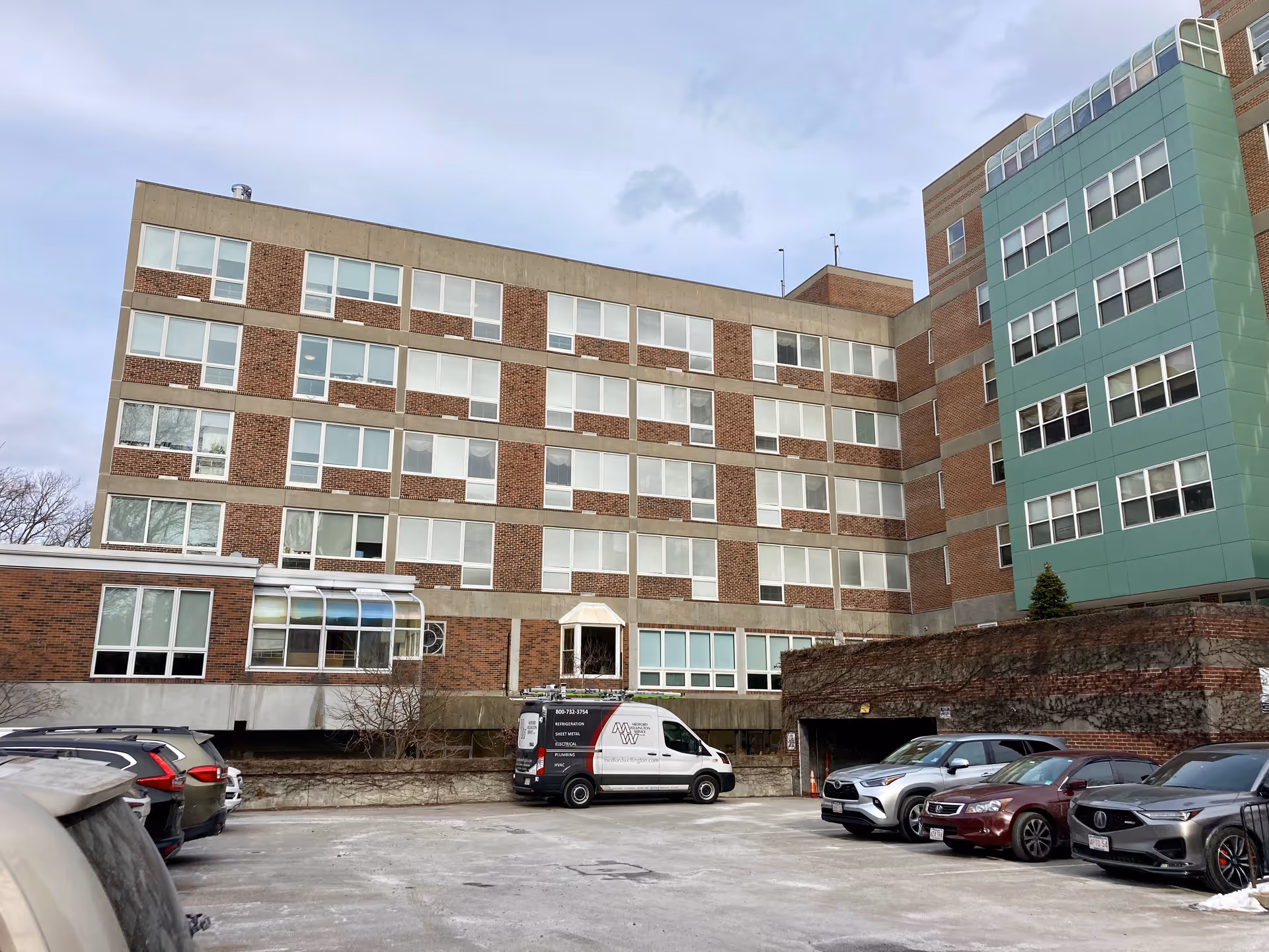 Exterior view of a multi-story brick and concrete building with many windows, adjacent to a parking lot with several parked cars and a white service van. The sky is overcast.