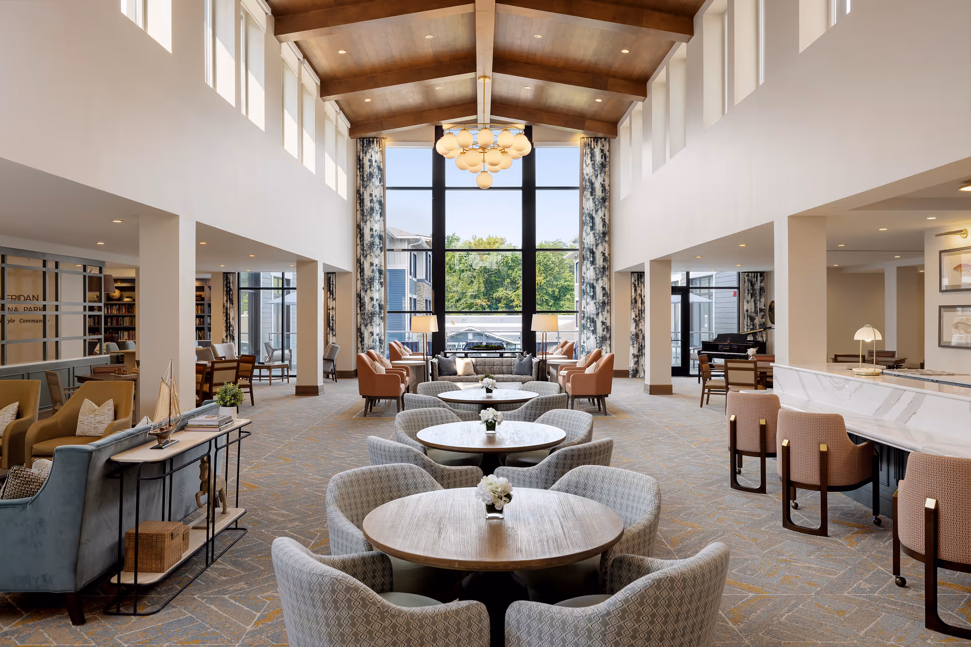 Bright senior living lobby with round tables, plush seating, and a high wooden ceiling.