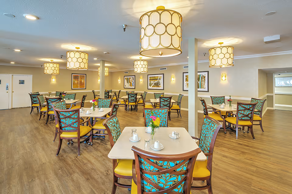 A bright and spacious dining room in a senior living facility with multiple tables and chairs. The chairs have wooden frames with turquoise and yellow patterned upholstery. Each table is set with white cups, saucers, and small vases containing flowers. The room features wooden flooring, beige walls adorned with framed artwork, and several large, decorative ceiling lights.