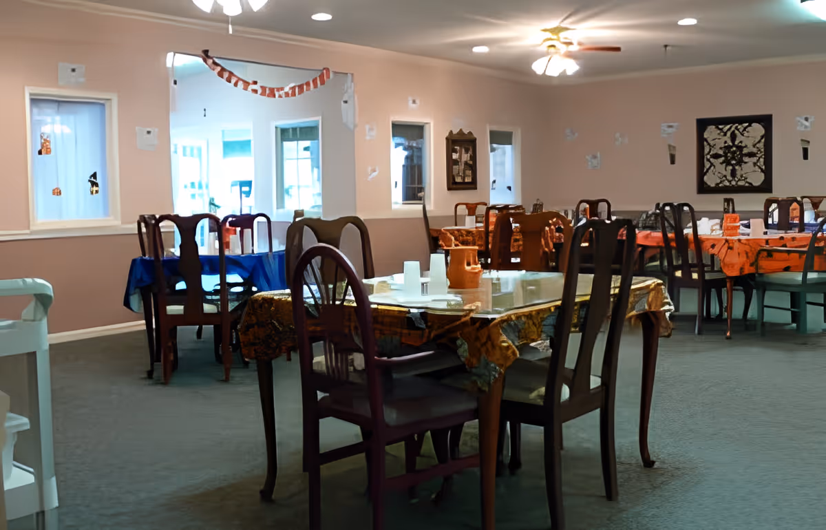 Communal dining room with multiple tables and wooden chairs, each table covered with tablecloths and place settings.