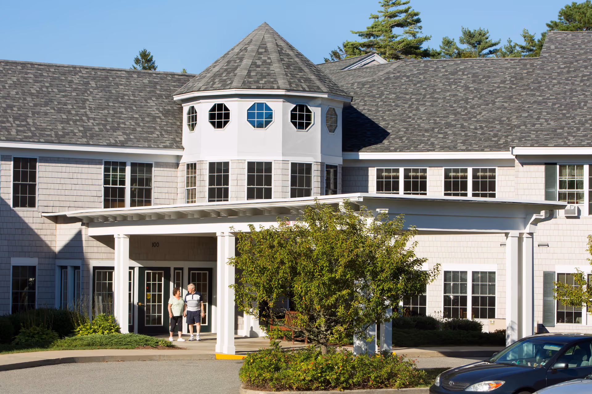 Front entrance of a multi-story senior living building with a covered portico, octagonal tower windows, landscaping, and two people near the doorway.