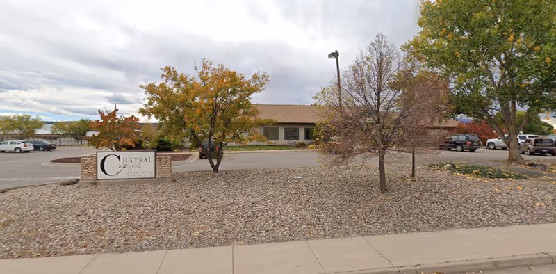 Exterior view of Chateau At Rifle facility showing a single-story building with a beige roof, surrounded by a parking lot with several parked cars. In the foreground, there is a landscaped area with rocks, small trees with autumn foliage, and a sign that reads 'Chateau At Rifle'. The sky is cloudy.
