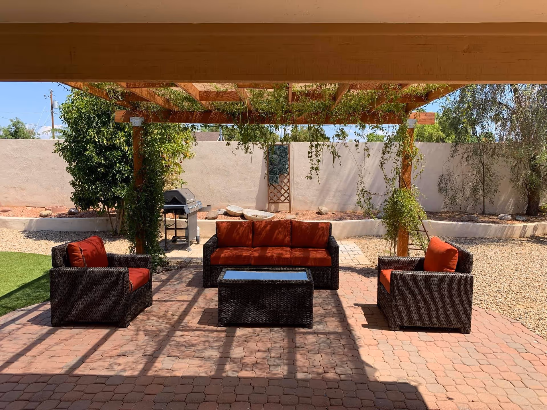 Outdoor patio area with a wooden pergola covered in hanging vines. Underneath, there is a seating arrangement consisting of a wicker sofa and two armchairs with red cushions, and a matching wicker coffee table with a glass top. In the background, there is a barbecue grill, some trees, and a beige stucco wall enclosing the space.