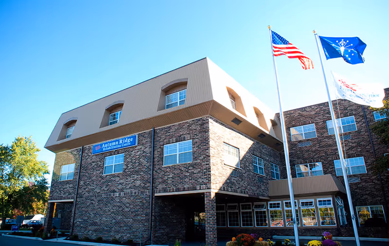 Exterior view of Autumn Ridge Rehabilitation Centre, a multi-story brick building with several windows. Three flagpoles stand in front of the building, displaying the American flag, a blue flag with a torch symbol, and another flag with text. Trees and a clear blue sky are visible in the background.