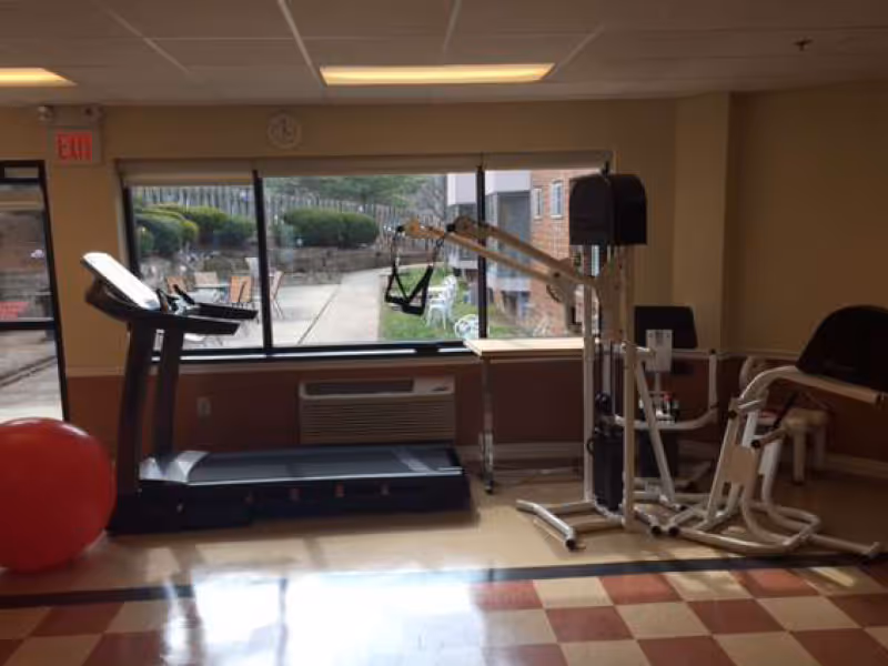 Interior fitness room with treadmill, exercise machines, red stability ball by windows.