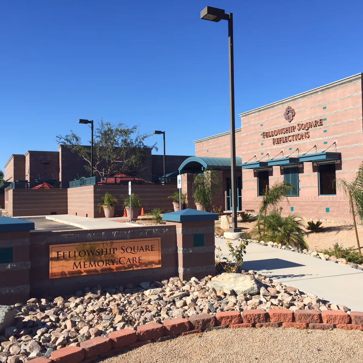 Front exterior of a reddish-beige brick senior living facility with a 'Fellowship Square Memory Care' sign, walkway, and rock landscaping.