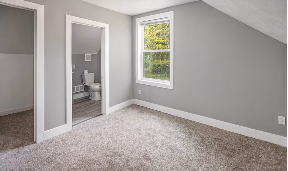 Empty gray-walled room with carpet, a doorway to a bathroom, and a window overlooking trees.