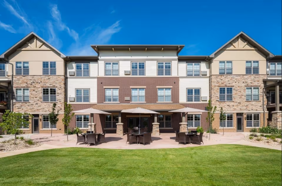 Exterior view of a three-story retirement living building with a mix of brick and stone facade, large windows, and a patio area with tables, chairs, and umbrellas set on a paved surface, surrounded by green grass and small trees under a blue sky.