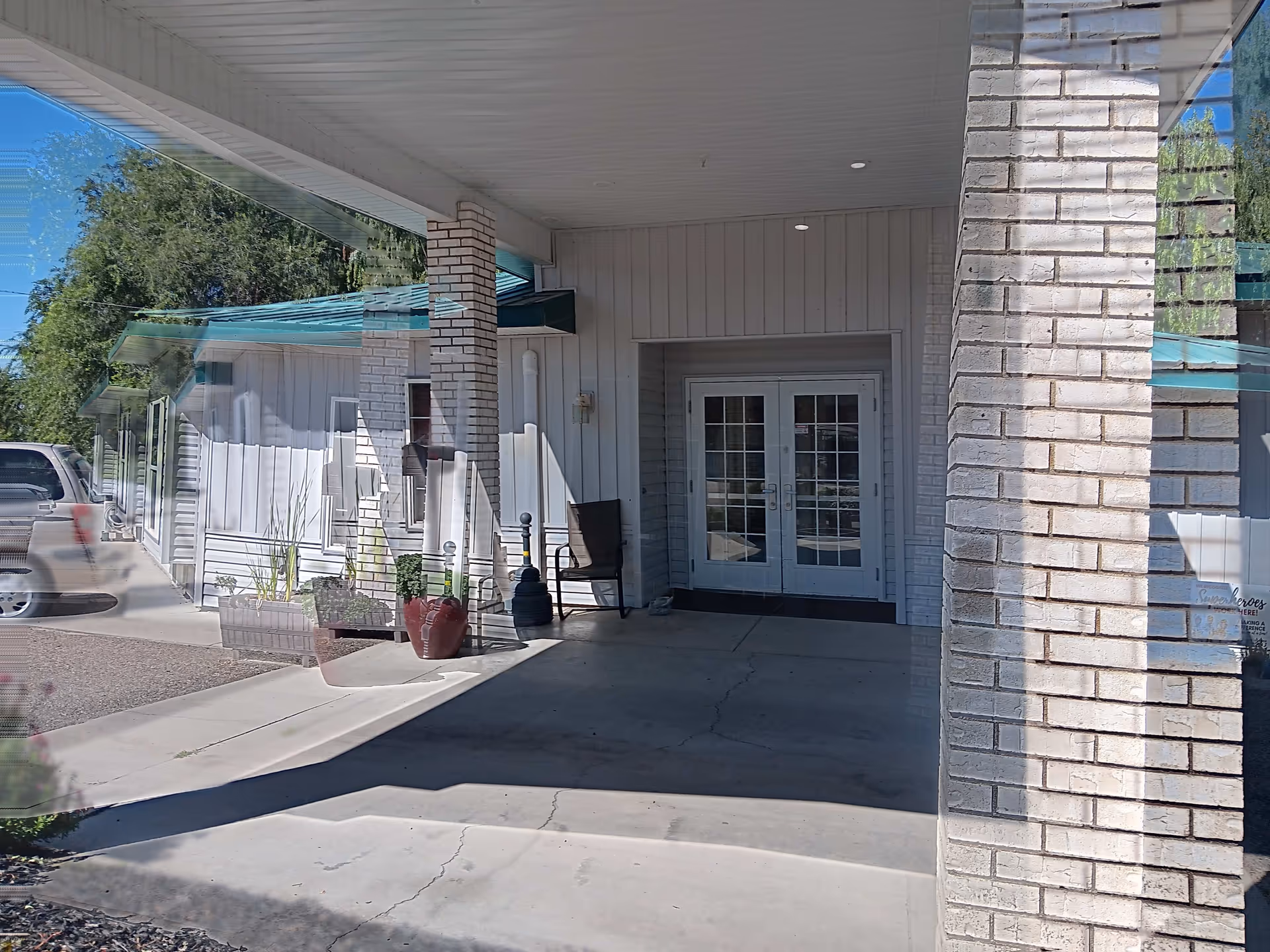 Covered entrance area of a building with white brick pillars and double glass doors. There is a chair and some potted plants near the entrance. A vehicle is partially visible on the left side, and the building has white siding with a teal roof.