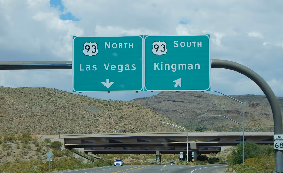 Two large green highway signs over a desert highway with mountains in the background.