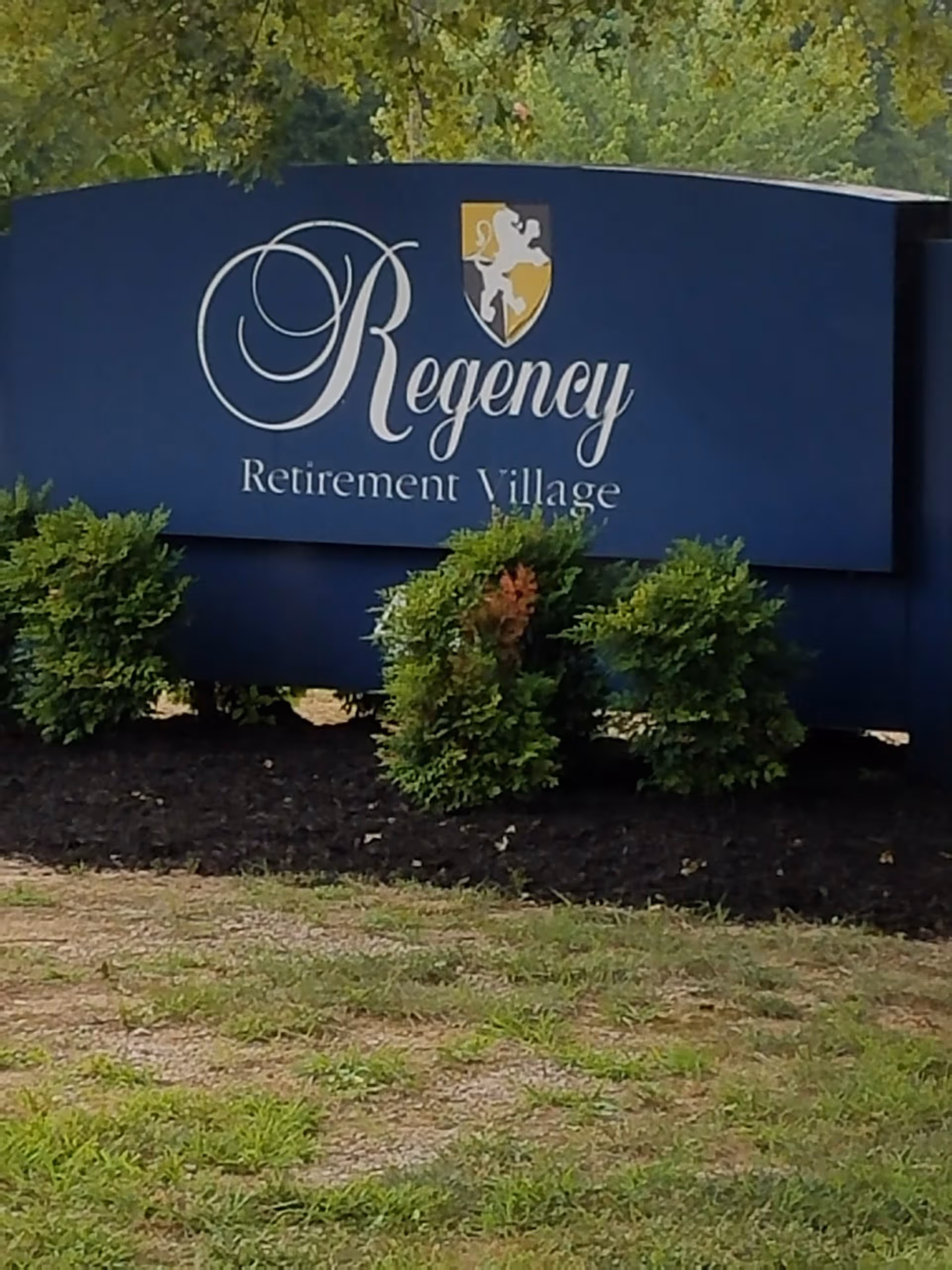 Large blue Regency Retirement Village sign with shrubs and trees in front of the facility