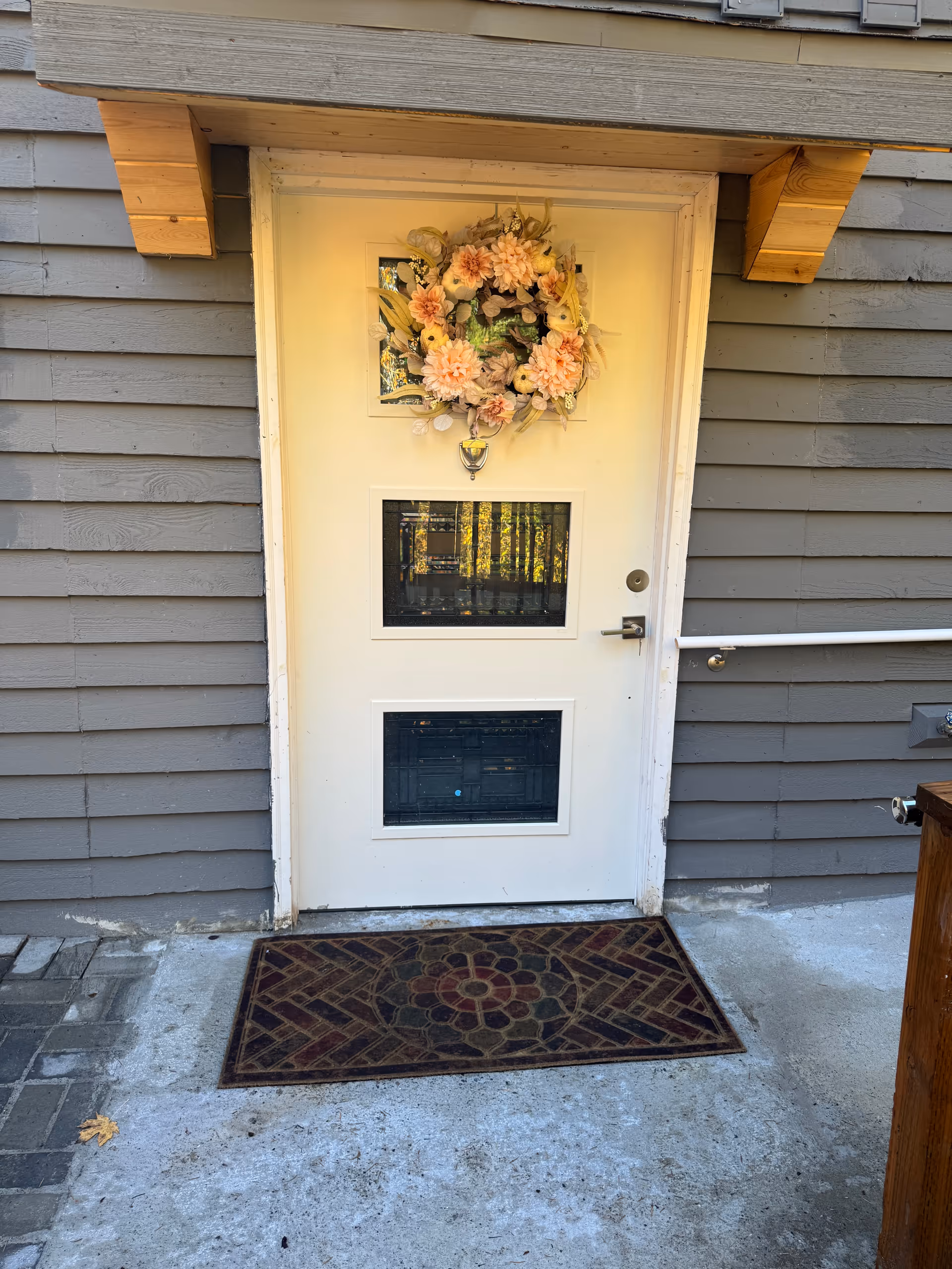 White front door with a wreath, gray siding, and a patterned mat.