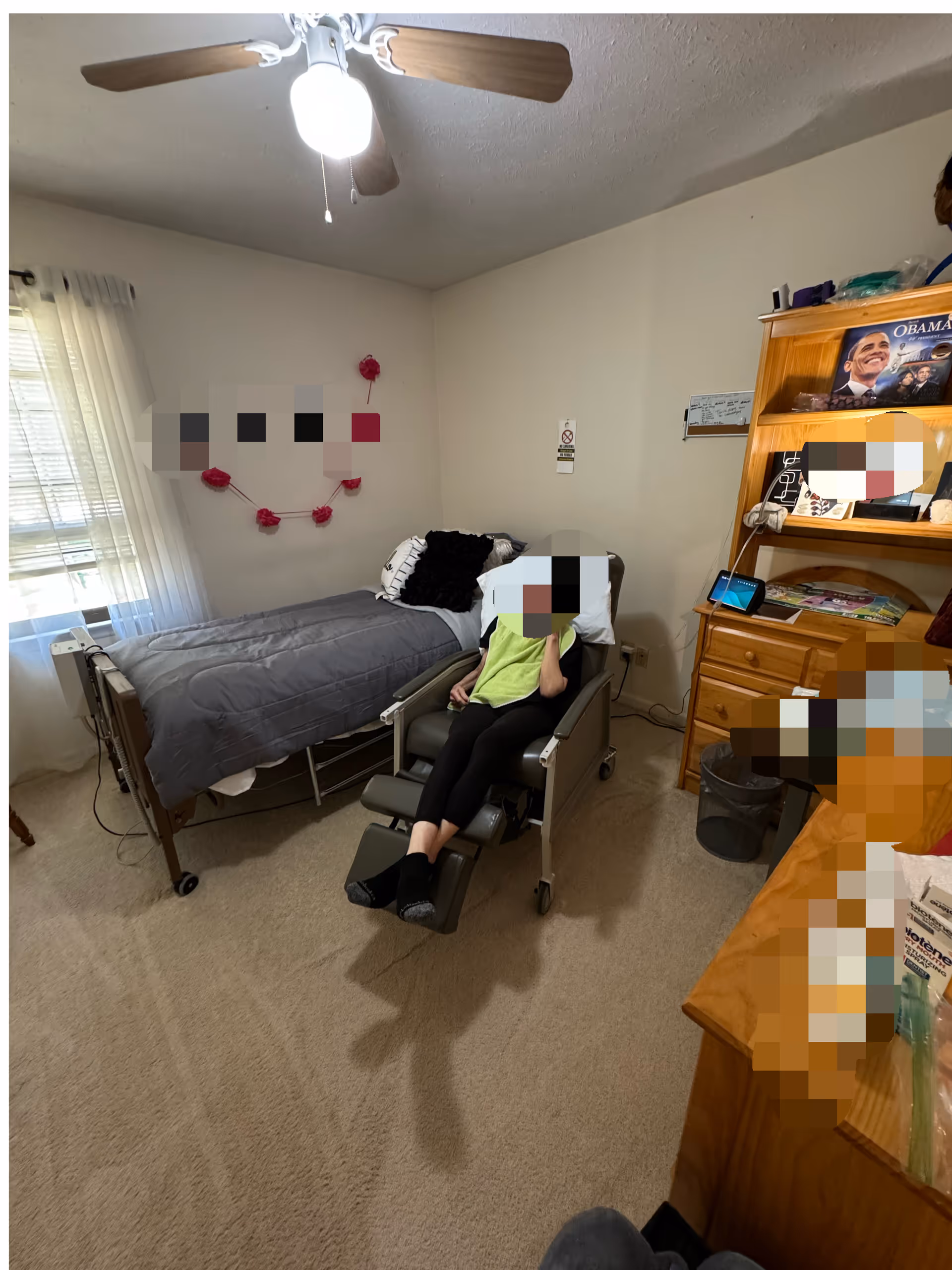 A resident sits in a reclining chair beside a bed in a small, well-lit bedroom.