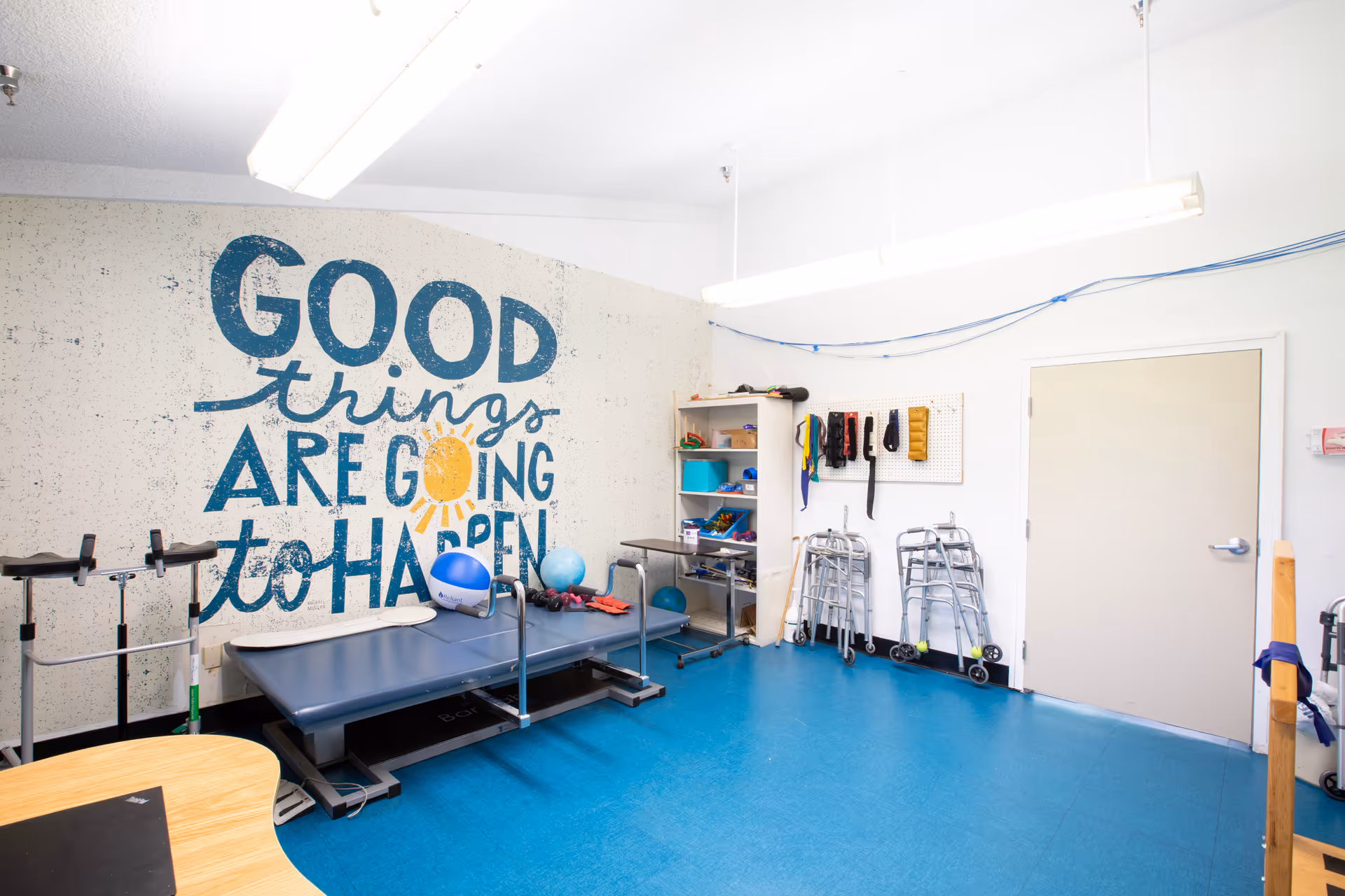 Bright therapy/exercise room with blue floor, equipment, walkers and a mural reading "GOOD things ARE GOING to HAPPEN" on the wall.