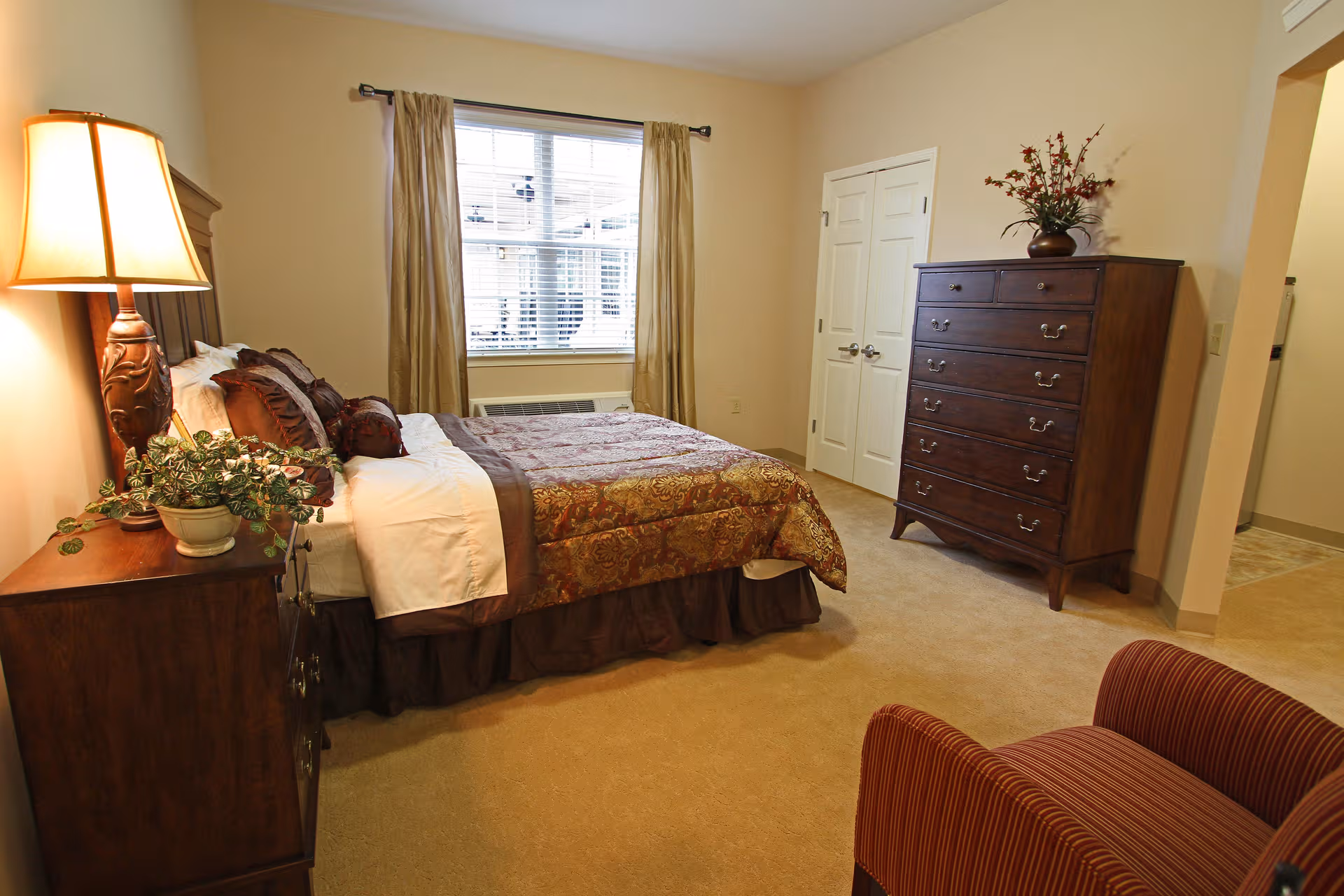 A well-appointed bedroom in a senior living facility featuring a large bed, dresser, and seating.