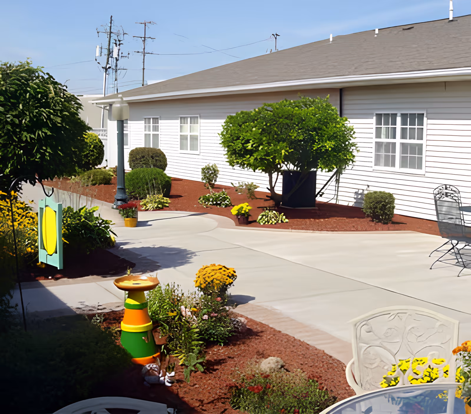 Outdoor patio area at Camillus Ridge Terrace featuring a concrete walkway, landscaped garden beds with various shrubs and flowers, a colorful birdbath, and metal patio chairs and tables. The building exterior is white with multiple windows and a gray roof under a clear blue sky.