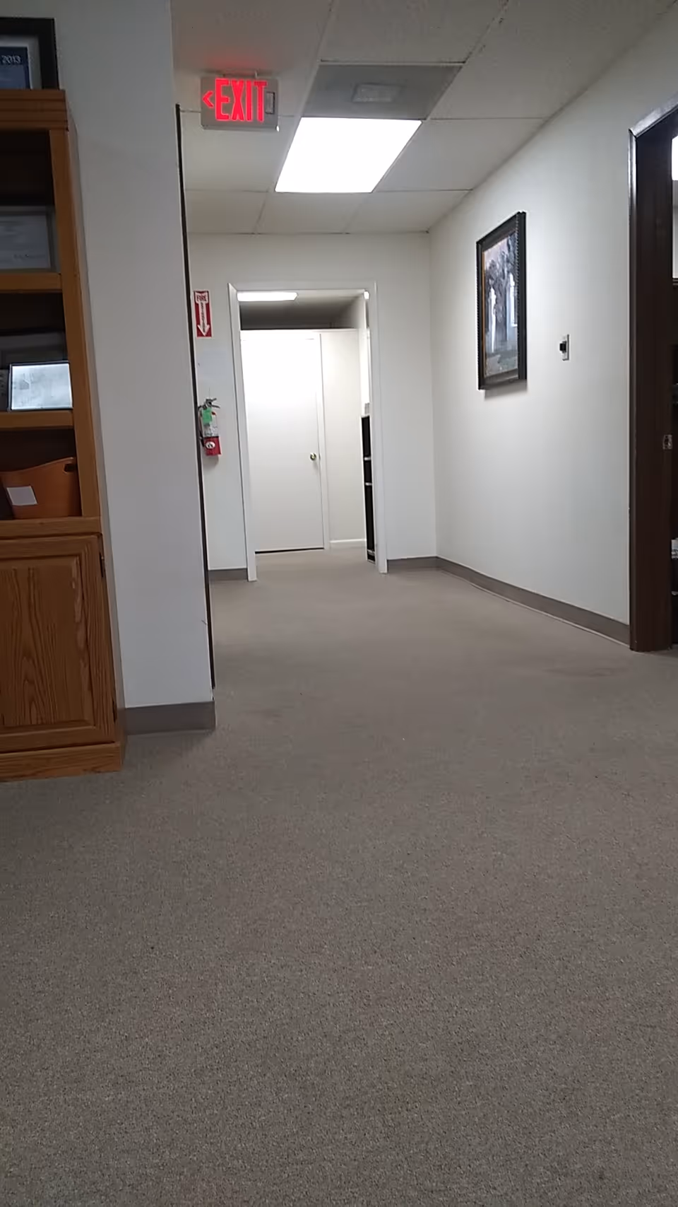 Long hallway inside a facility with ceiling lights, an exit sign, and framed wall art.