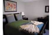 Bedroom in a senior living facility with a green bedspread, pillows, lamps, and floral wall art.