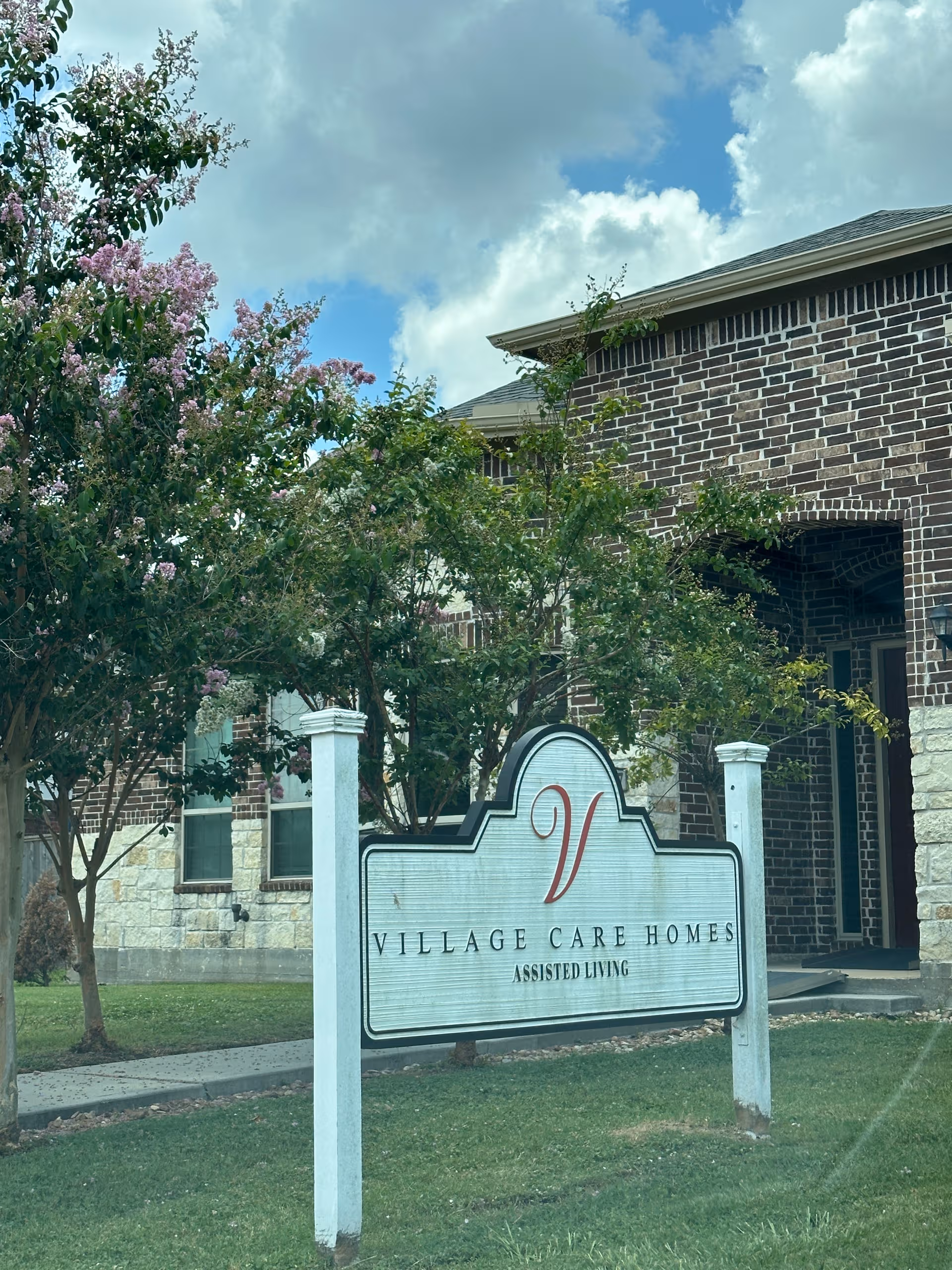 Front view of Village Care Homes Assisted Living sign beside brick building and trees.